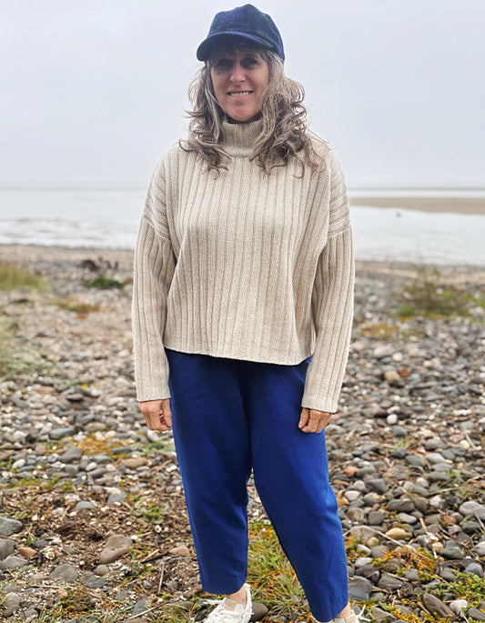 A woman standing on a rocky beach wearing a cream ribbed turtleneck sweater, blue pants, a navy cap, and white sneakers.