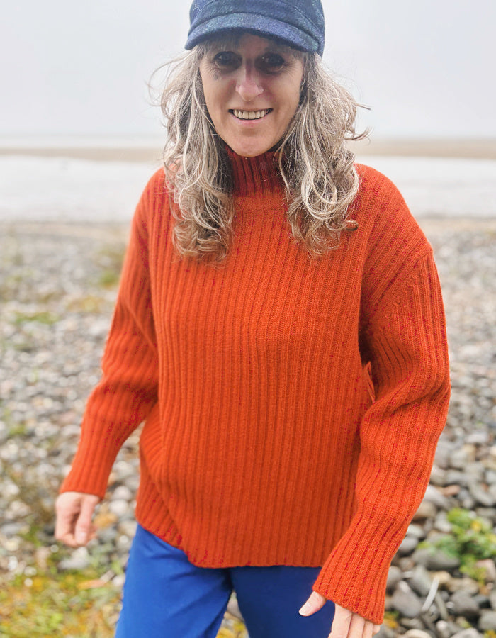 A woman wearing a ribbed orange turtleneck sweater, blue pants, and a blue cap, standing outdoors on a pebble beach.