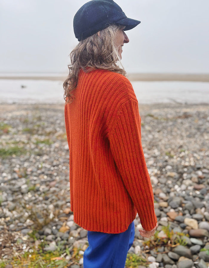 A person standing on a pebble beach wearing a bright orange ribbed knit sweater, blue pants, and a dark navy baseball cap, photographed from the side/back in an outdoor coastal setting.