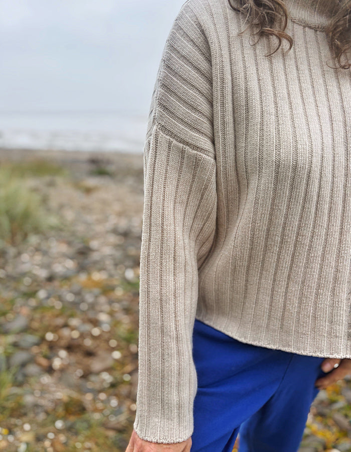 A close-up of a person wearing a beige ribbed knit sweater and bright blue pants, standing outdoors on a rocky beach with a blurred natural background.