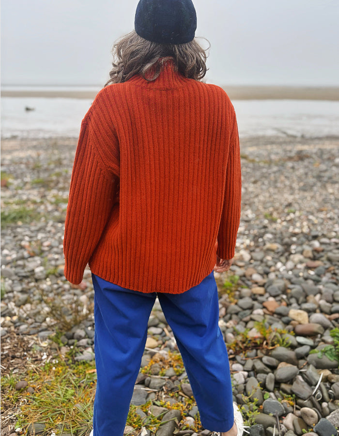 A person standing on a rocky beach, viewed from behind, wearing a ribbed orange-red turtleneck sweater, blue pants, and a black beanie.