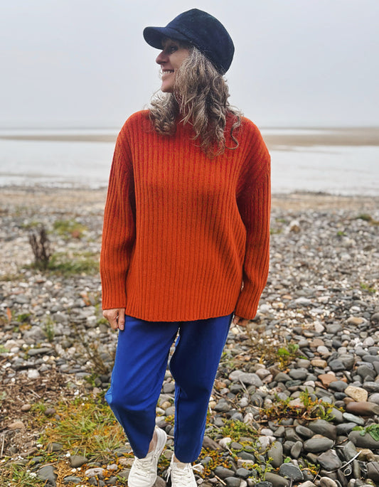A woman standing on a rocky beach wearing a chunky orange knit sweater, blue pants, white sneakers, and a black cap, looking to the side.