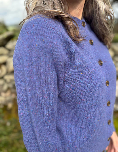 Woman wearing a purple donegal tweed knitted cardigan and patterned hat standing in a natural setting with trees and a stone wall.