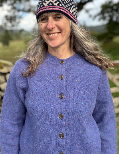 Woman wearing a purple donegal tweed knitted cardigan and patterned hat standing in a natural setting with trees and a stone wall.