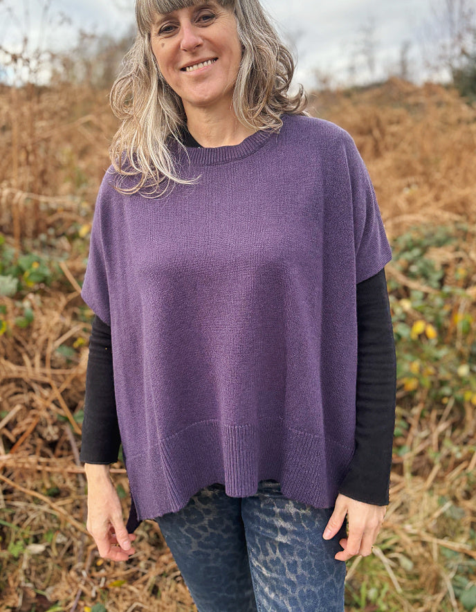 Woman wearing a purple poncho in an outdoor setting with dry grass and trees.