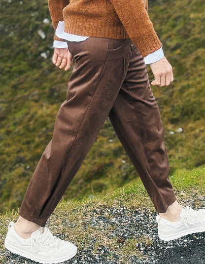 A person walking outdoors wearing brown tapered trousers, a rust-orange knit sweater, a white shirt, and white sneakers, with a grassy hill in the background.