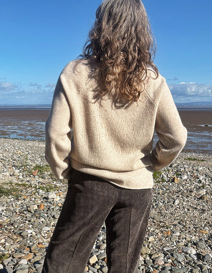 A person standing on a pebble beach facing the ocean, wearing a cream-colored ribbed knit sweater and dark brown corduroy pants.