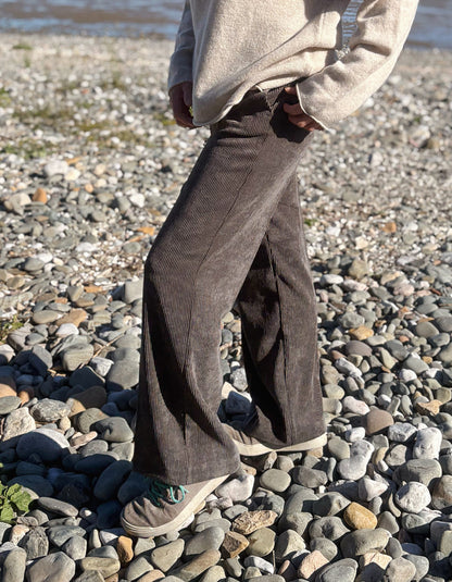 A person standing on a pebble beach wearing dark brown corduroy wide-leg trousers, a beige knit sweater, and gray sneakers.