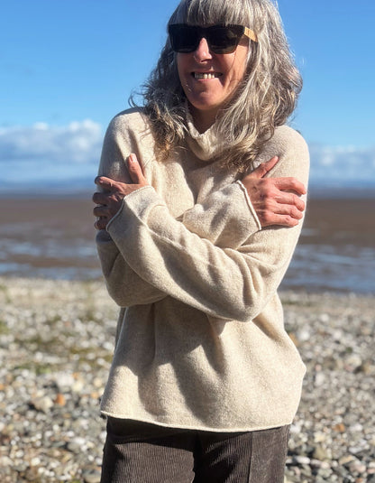 A woman with gray hair and sunglasses stands on a pebble beach with her arms crossed, wearing a cream-colored knit turtleneck sweater.