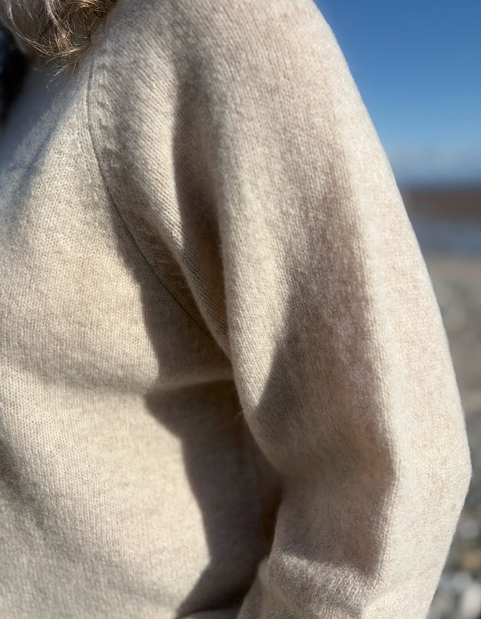 Close-up of a person wearing a soft, light beige knit sweater outdoors, with sunlight casting shadows across the fabric and a blurred blue sky and shoreline in the background.