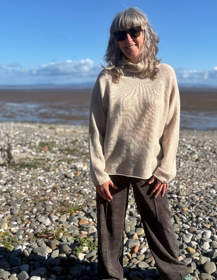 A woman with gray hair and sunglasses stands on a pebble beach wearing a cream-colored ribbed turtleneck sweater and dark brown wide-leg trousers, with water and a blue sky in the background.