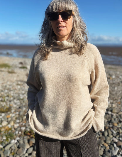 A woman with gray hair and sunglasses wearing a beige turtleneck sweater, standing on a rocky beach with water and sky in the background.