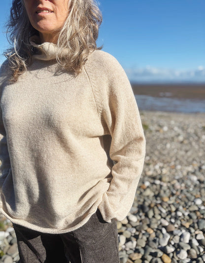 A woman wearing a light beige knit turtleneck sweater stands on a pebble beach with water and blue sky in the background.