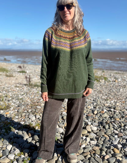 A woman standing on a rocky beach wearing a dark green knit sweater with a multicolor geometric pattern across the chest, brown corduroy pants, and sunglasses.