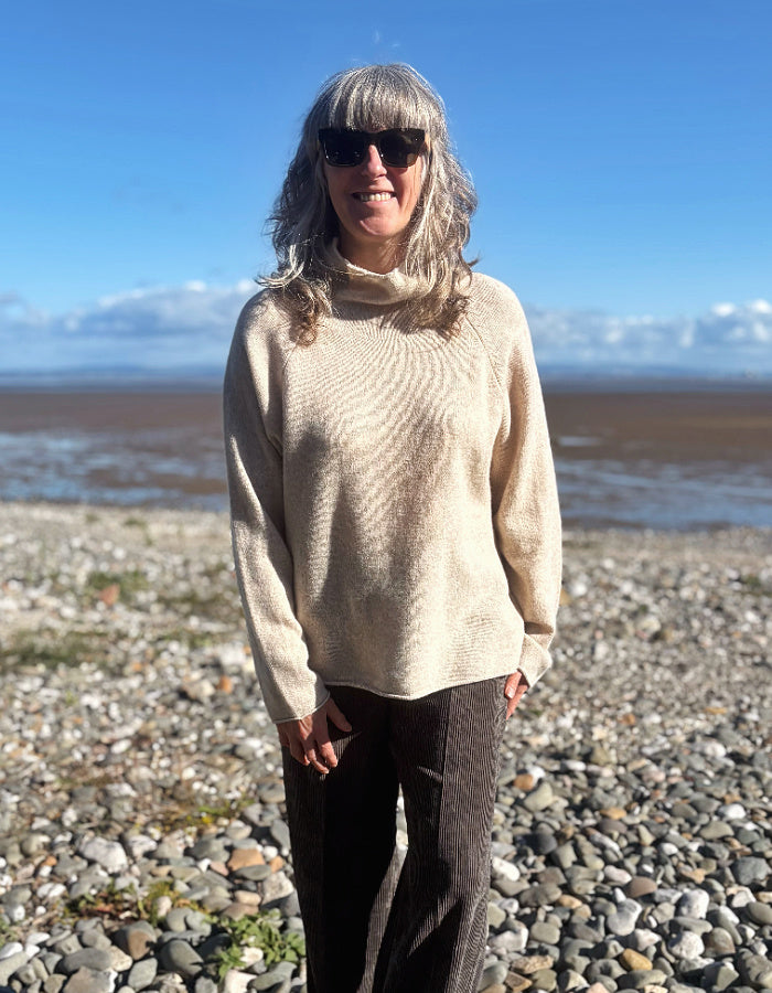 A woman with gray hair and sunglasses stands on a pebble beach wearing a cream-colored knit turtleneck sweater and dark pants, with water and mountains in the background under a blue sky.