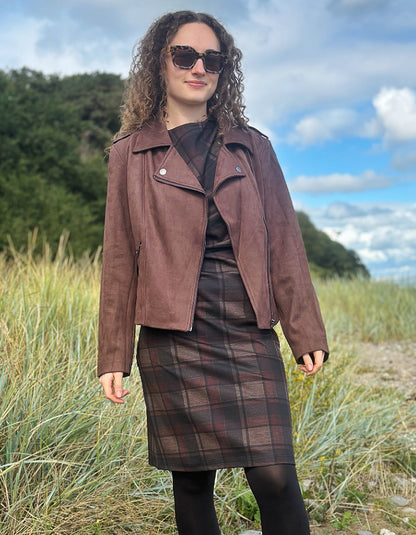 A woman wearing a brown faux-leather moto jacket, a dark plaid skirt, and sunglasses, standing outdoors in a grassy field under a cloudy blue sky.