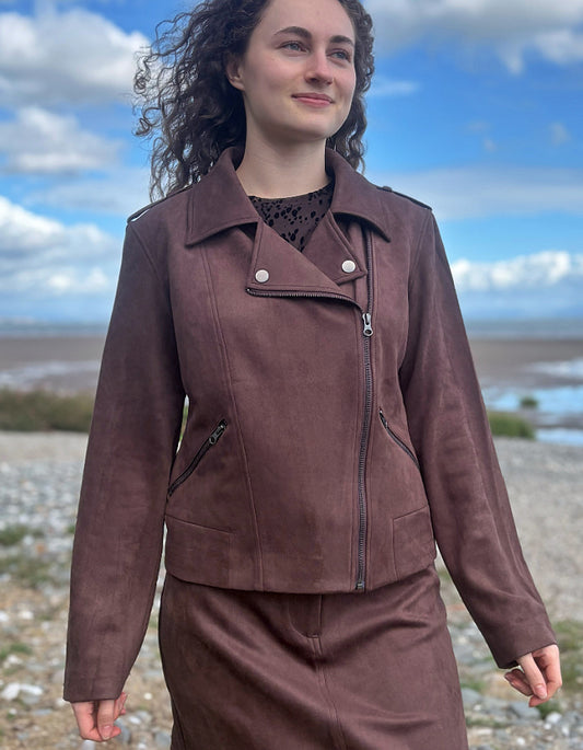 A woman wearing a brown faux-leather moto jacket and matching skirt stands outdoors on a pebble beach with water and a cloudy blue sky in the background.