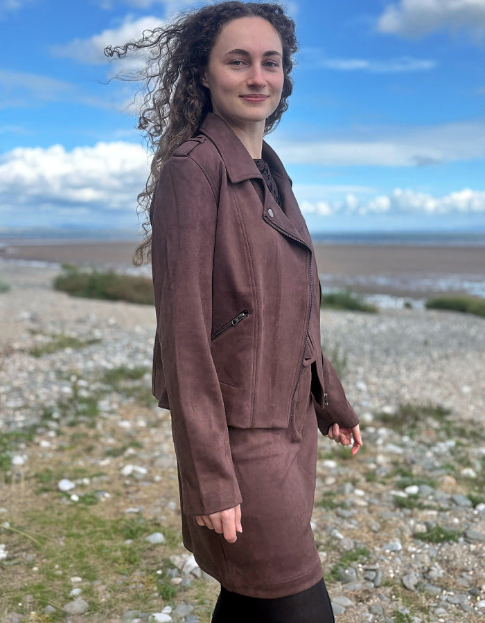 A woman with long curly brown hair stands outdoors on a rocky shoreline under a blue sky, wearing a brown suede-like jacket and matching skirt.