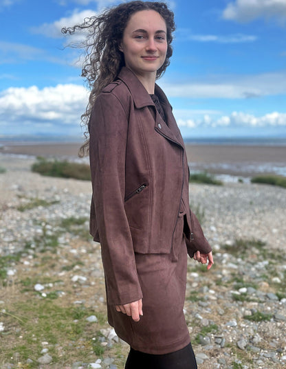 A woman with long curly brown hair stands outdoors on a rocky shoreline under a blue sky, wearing a brown suede-like jacket and matching skirt.