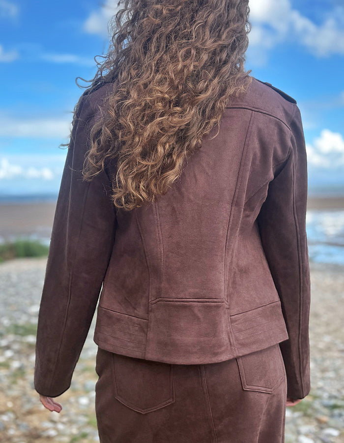 A person viewed from behind wearing a brown suede-like jacket and matching skirt set, standing outdoors on a pebble beach with water and blue sky in the background.