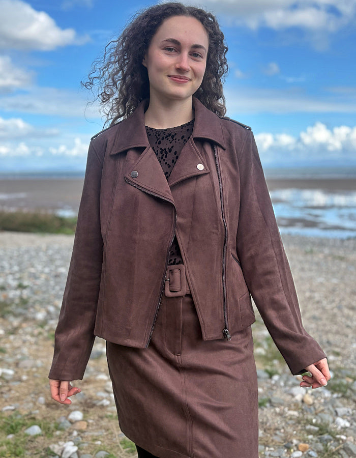 A woman with curly brown hair stands outdoors on a pebble beach wearing a matching brown faux-leather moto jacket and skirt set over a black lace top, with a blue sky and water in the background.
