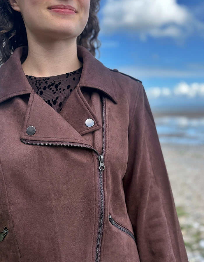 A close-up of a person wearing a brown suede moto jacket with an asymmetrical zipper and snap button, set against a blurred beach and sky background.