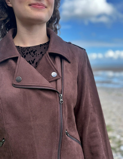 A close-up of a person wearing a brown suede moto jacket with an asymmetrical zipper and snap button, set against a blurred beach and sky background.