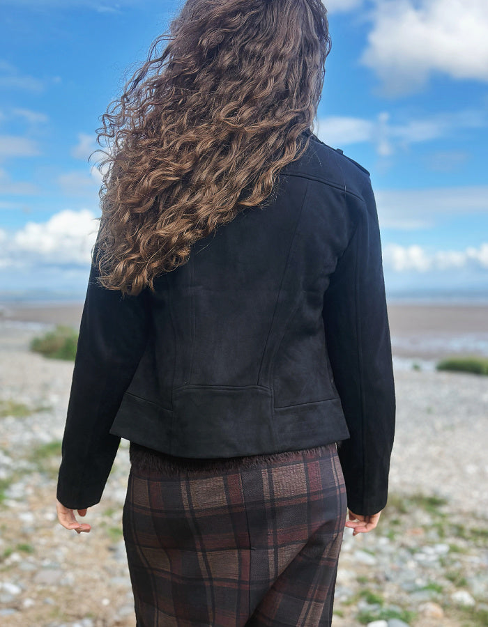 A person viewed from behind wearing a black cropped jacket and brown plaid pants, standing outdoors on a rocky shoreline under a blue sky with clouds.