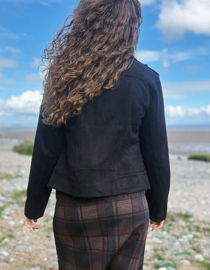 A person viewed from behind wearing a black cropped jacket and brown plaid pants, standing outdoors on a rocky shoreline under a blue sky with clouds.