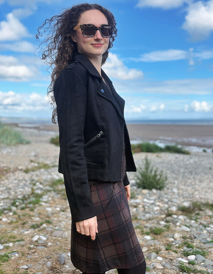 A woman wearing a black suede moto jacket and a brown plaid kilt stands on a rocky shoreline under a blue sky with clouds.