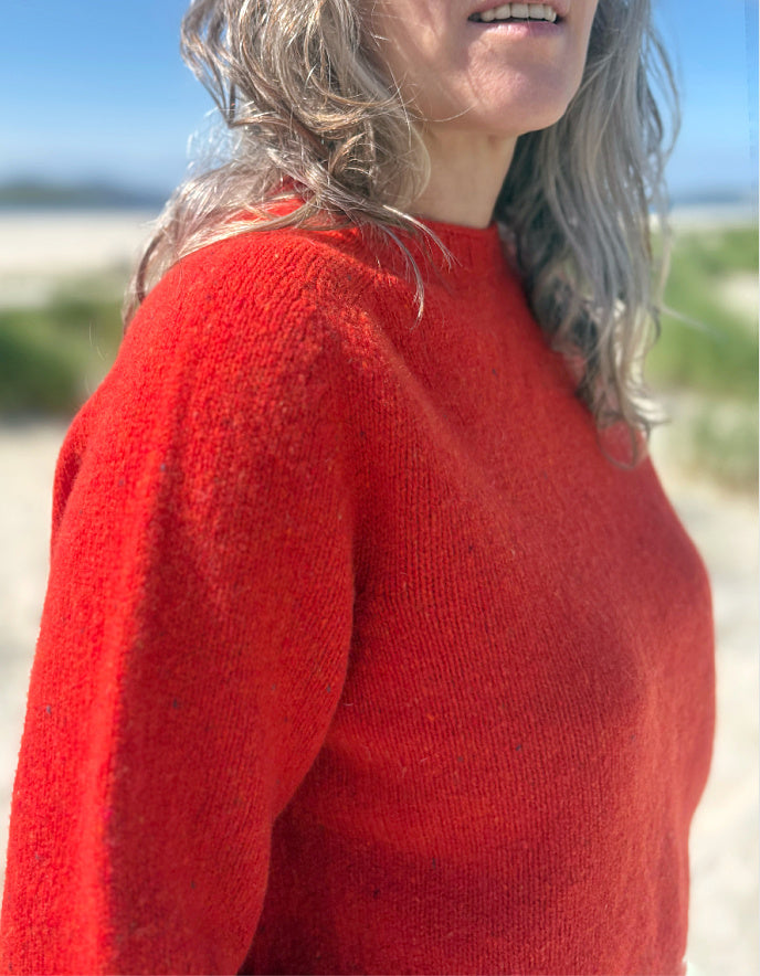 A close-up of a woman wearing a bright red knit sweater outdoors, with her hair blowing in the wind against a blurred beach background.