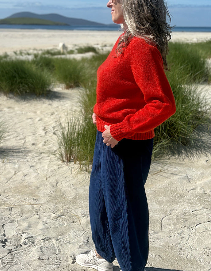A woman standing on a sandy beach wearing a bright red knit sweater and dark navy wide-leg pants, with dune grass and the ocean in the background.