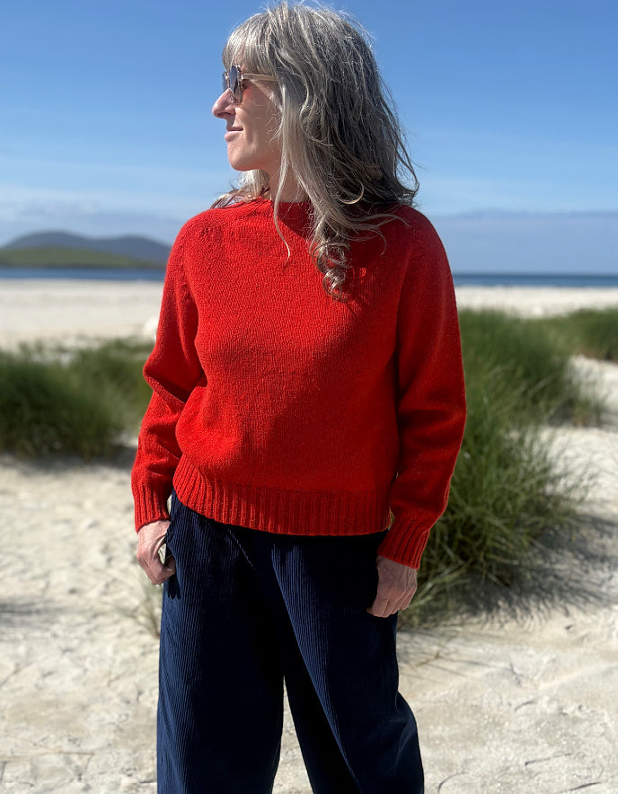 A woman standing on a sandy beach wearing a bright red knit sweater and dark navy pants, with blue sky and dune grass in the background.