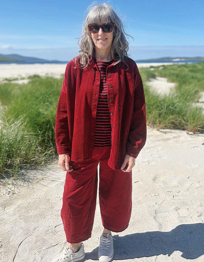 A woman stands outdoors on a sandy path near beach grass under a blue sky, wearing a matching red corduroy jacket and cropped pants over a striped shirt and white sneakers.
