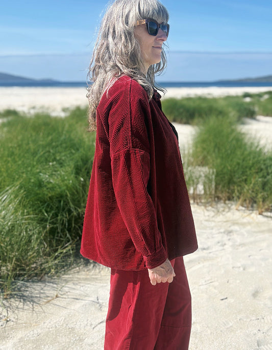 A woman with gray hair and sunglasses stands on a sandy beach wearing a matching red corduroy long-sleeve top and pants set, with green beach grass and blue sky in the background.