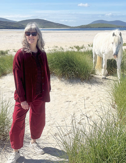 A woman wearing a matching burgundy corduroy jacket and trousers set stands on a sandy beach path with tall green grass, a white horse in the background, and mountains under a blue sky.