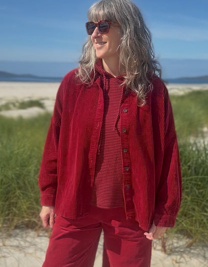 A woman wearing a matching red corduroy button-up shirt and pants set, layered over a red striped top, standing on a sandy beach with dune grass and blue sky in the background.