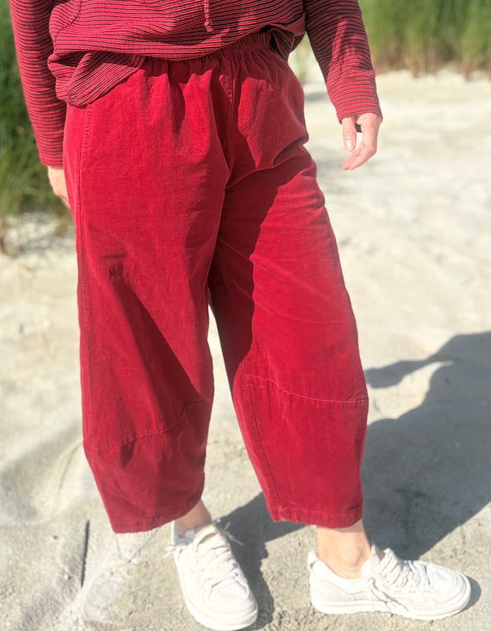 A person standing outdoors on sand wearing loose red corduroy cropped barrel pants, a red striped long-sleeve top, and white sneakers.