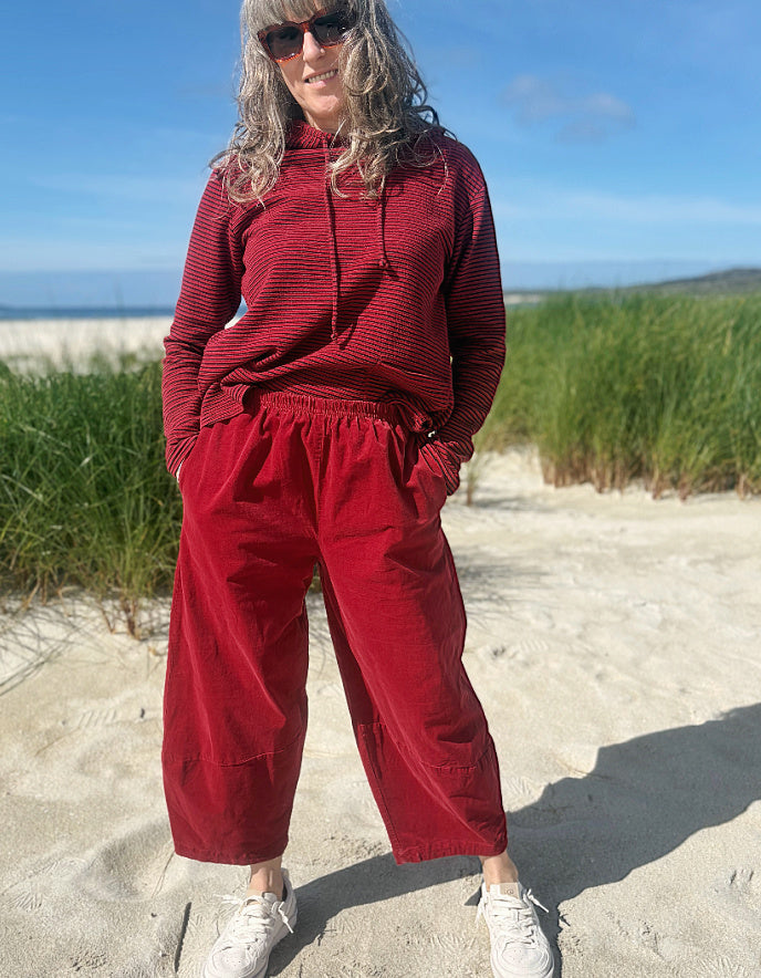 A woman stands on a sandy beach wearing a matching red striped long-sleeve top and red wide-leg pants, with sunglasses and white sneakers.
