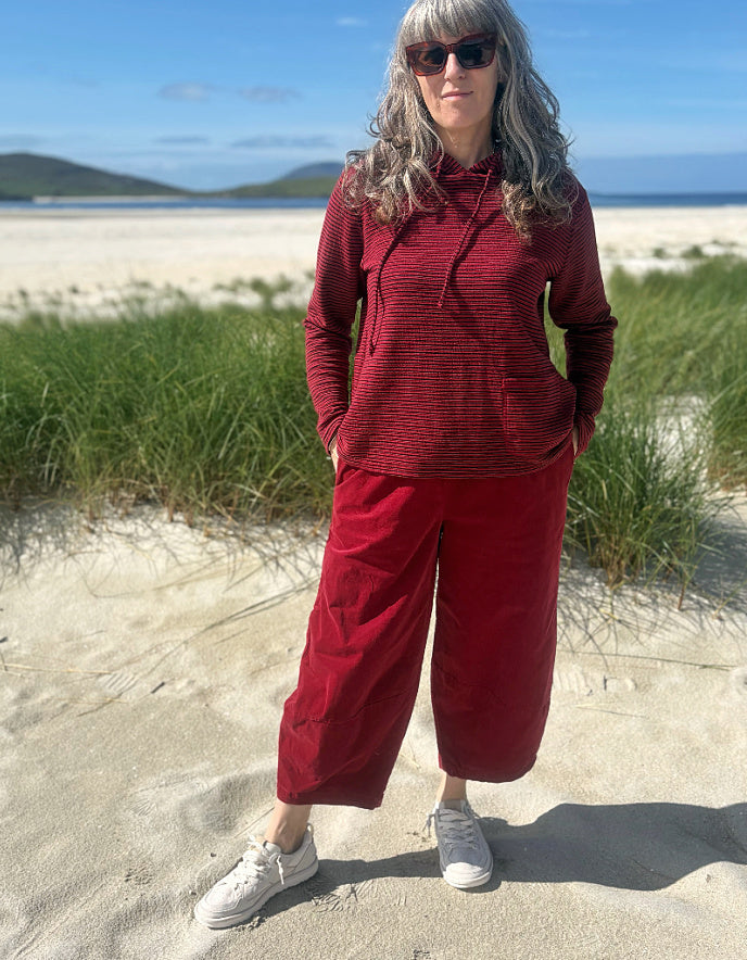 A woman stands on a sandy beach wearing a matching red ribbed knit hoodie and wide-leg cropped pants, with white sneakers and sunglasses, against a background of green grass, ocean, and blue sky.