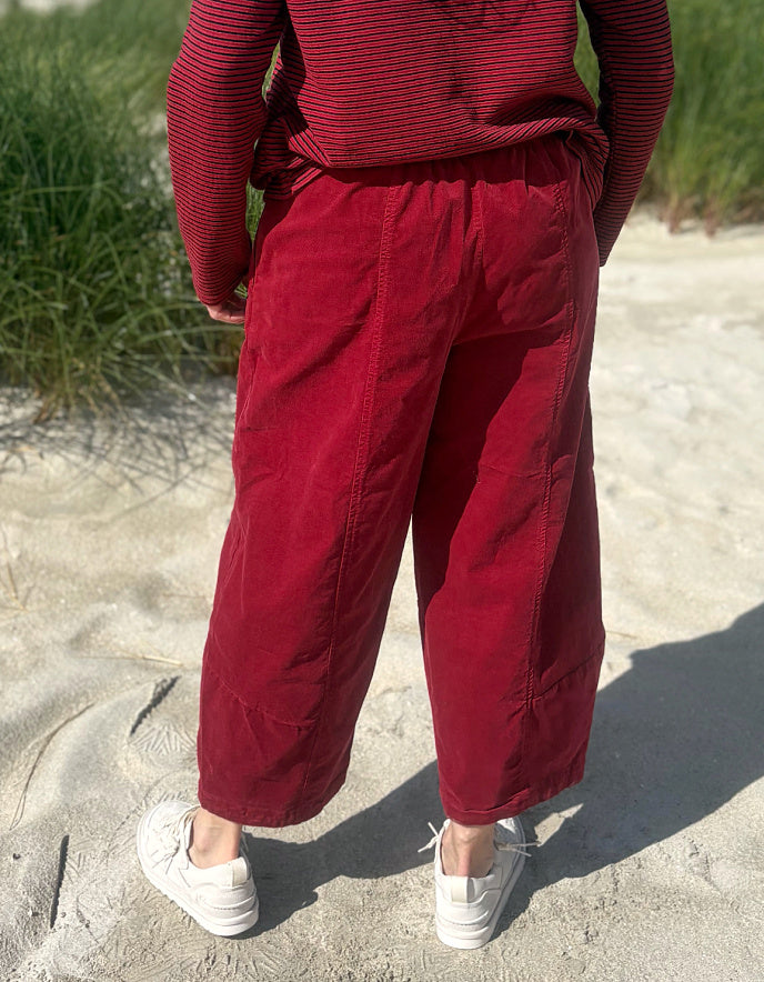 A person standing outdoors on sand wearing loose red cropped pants, a red striped long-sleeve top, and white sneakers, photographed from the back.