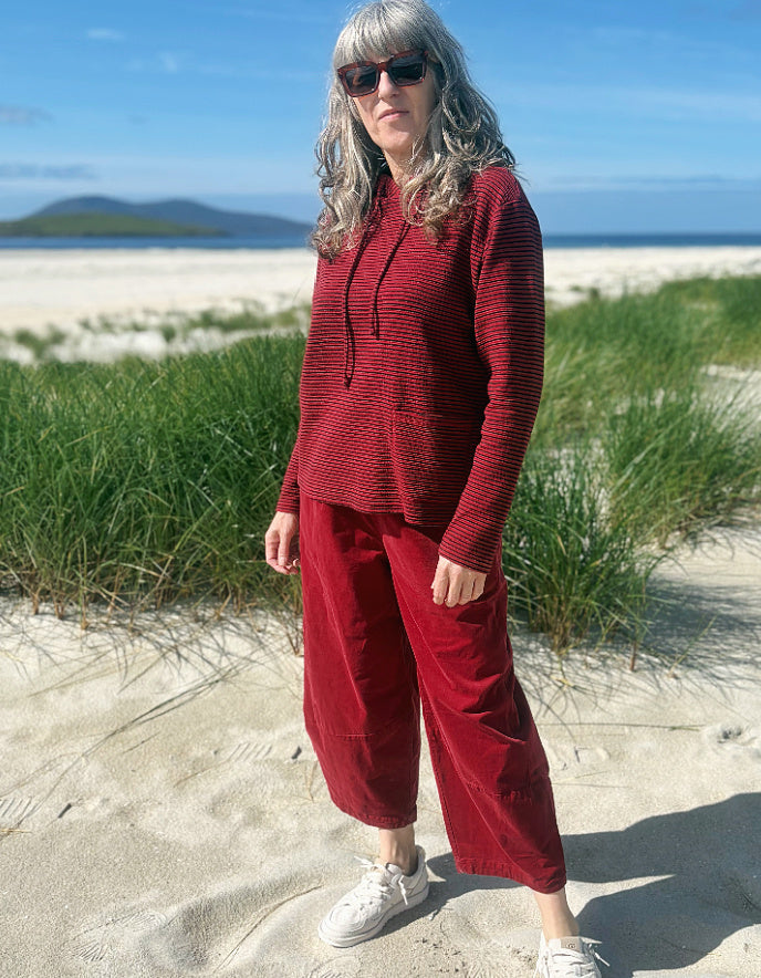 A woman wearing a matching red ribbed knit hoodie and wide-leg barrel cord pants set stands on a sandy beach with dune grass and the ocean in the background.