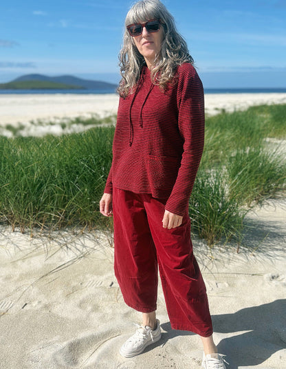 A woman wearing a matching red ribbed knit hoodie and wide-leg barrel cord pants set stands on a sandy beach with dune grass and the ocean in the background.