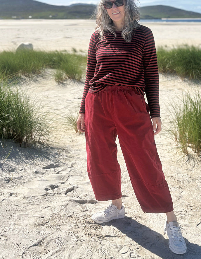A woman standing on a sandy beach dune wearing a red and black striped long-sleeve top, red wide-leg barrel cropped pants, and white sneakers.