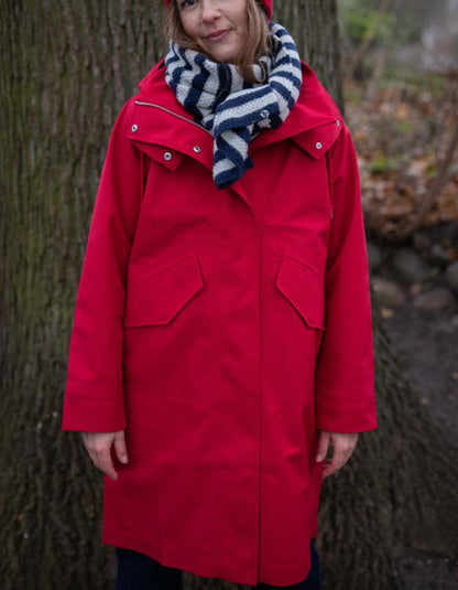 A woman wearing a bright red hooded coat with large front flap pockets, a navy and white striped knit scarf, and a red beanie, standing outdoors in front of a tree.