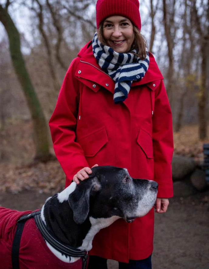 A woman wearing a bright red hooded coat, matching red knit beanie, and a navy-and-white striped scarf stands outdoors in a wooded area while petting a black-and-white dog wearing a red sweater.
