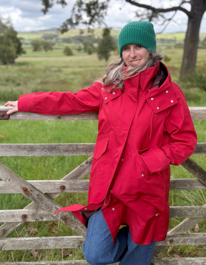 A woman wearing a bright red hooded parka and a green knit beanie leans against a rustic wooden fence in an outdoor grassy landscape.