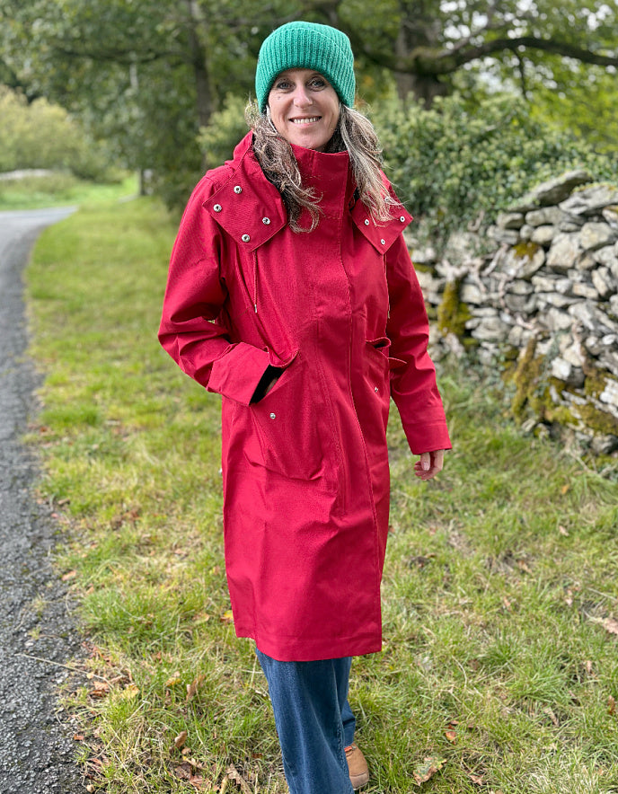 A woman wearing a bright red hooded parka jacket, blue jeans, and a green knit beanie stands outdoors on a grassy path next to a stone wall.
