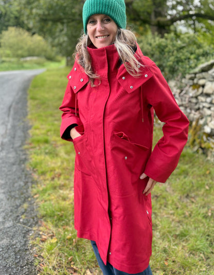 A woman wearing a bright red hooded raincoat and a green knit beanie stands on a country path outdoors.