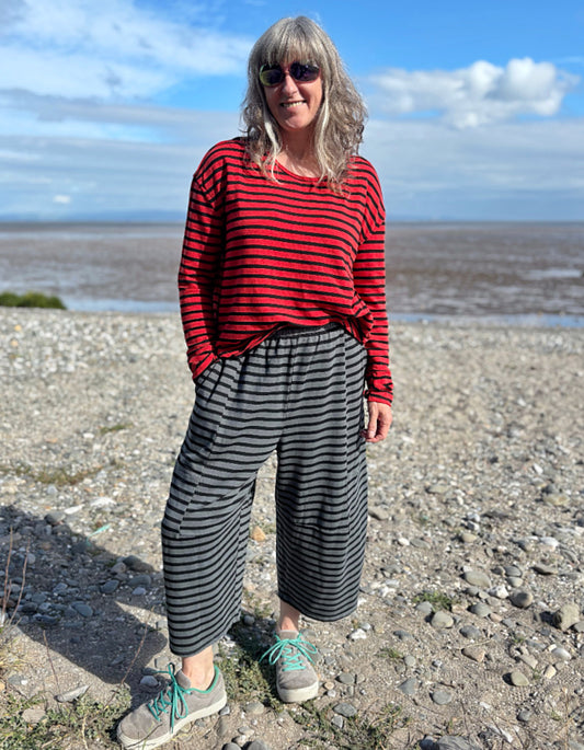 Person wearing a red and black striped shirt and black and gray striped pants standing on a pebbly beach.